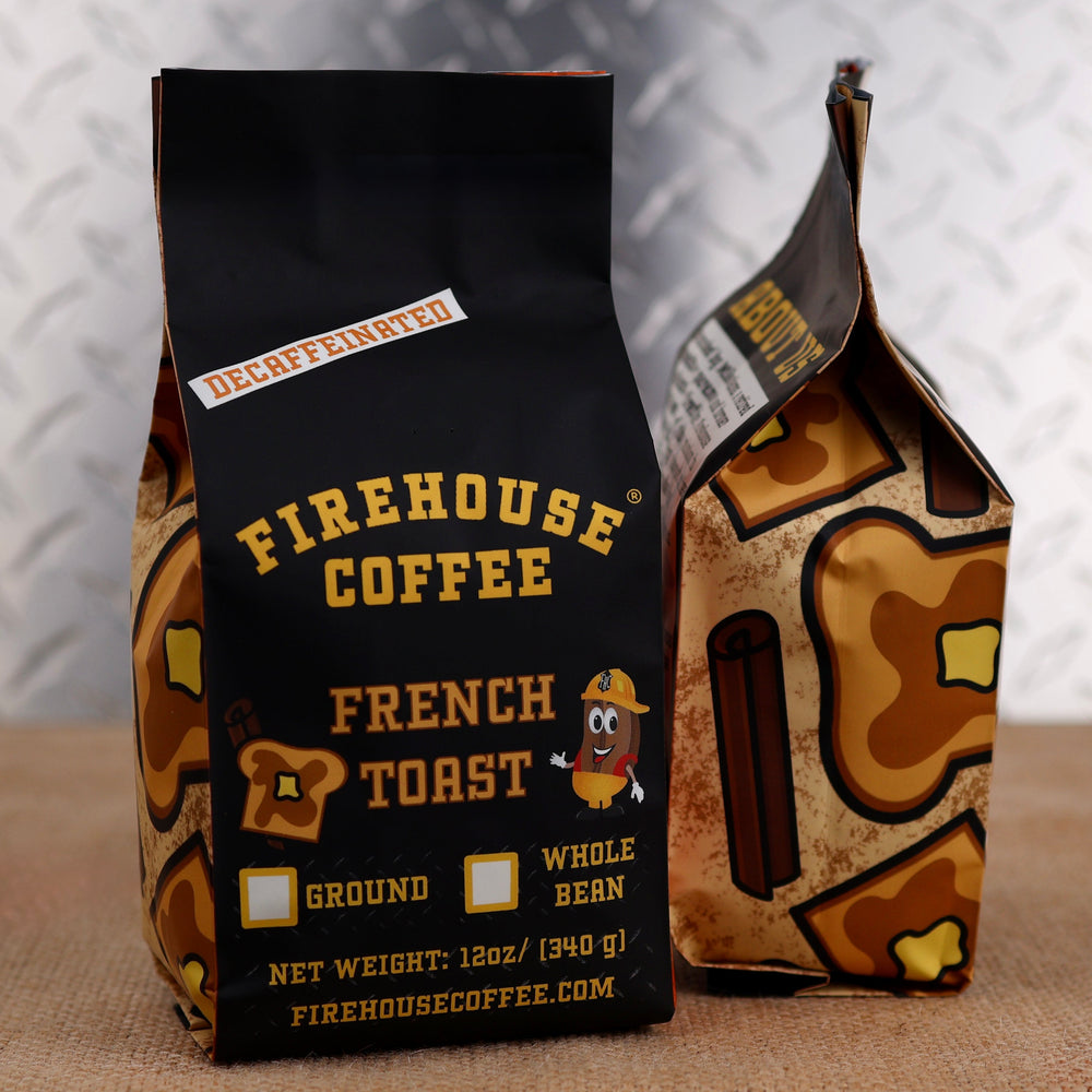 A black 12 oz Firehouse Coffee bag labeled French Toast decaffeinated coffee stands in front of a matching breakfast-themed patterned bag. The Swiss Water Process decaf coffee is available ground or whole bean and features Firehouse Coffee's signature firefighter branding.