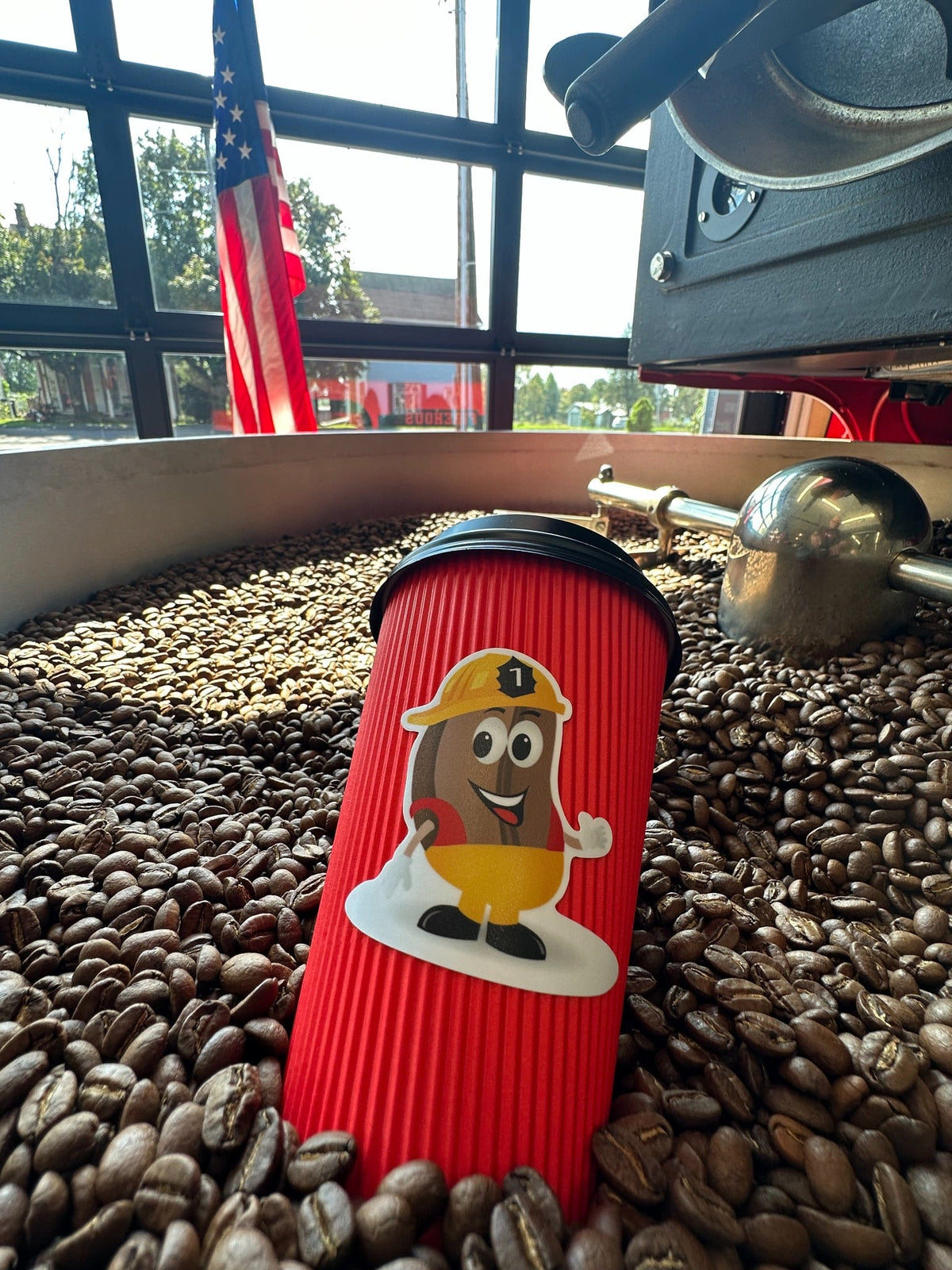 Red Firehouse Coffee mascot cup nestled in roasted coffee beans in a roaster cooling tray, with an American flag behind it.