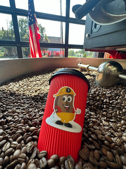 Red Firehouse Coffee mascot cup nestled in roasted coffee beans in a roaster cooling tray, with an American flag behind it.