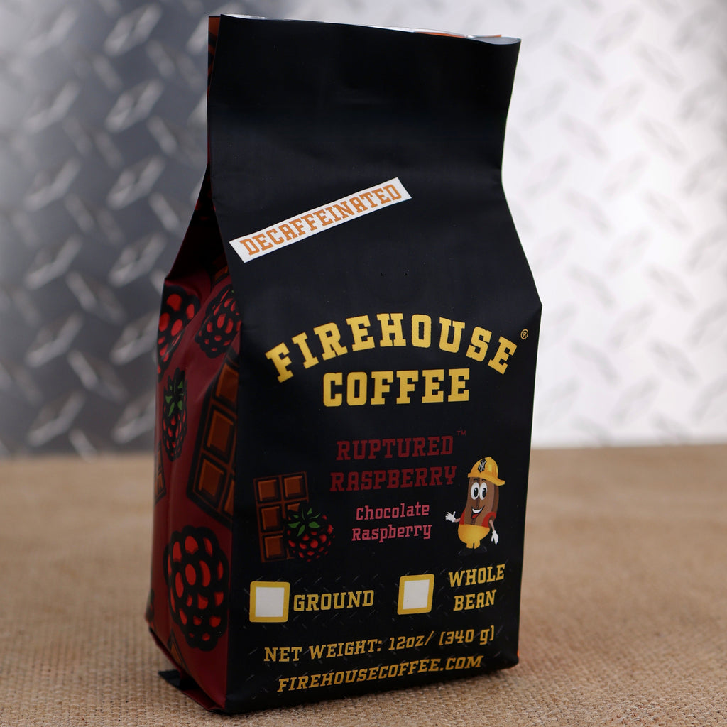 A black 12 oz Firehouse Coffee bag labeled Ruptured Raspberry stands in front of a raspberry and chocolate-themed patterned bag. The Swiss Water Process decaffeinated coffee features chocolate raspberry flavor and is available in ground or whole bean.