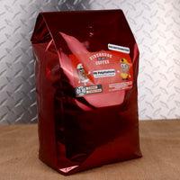A 5lb Bag of Firehouse Coffee bag labeled PB Palpitations stands in front of a red themed bag. The Swiss Water Process decaffeinated coffee features chocolate and peanut butter flavors and is available in ground or whole bean.