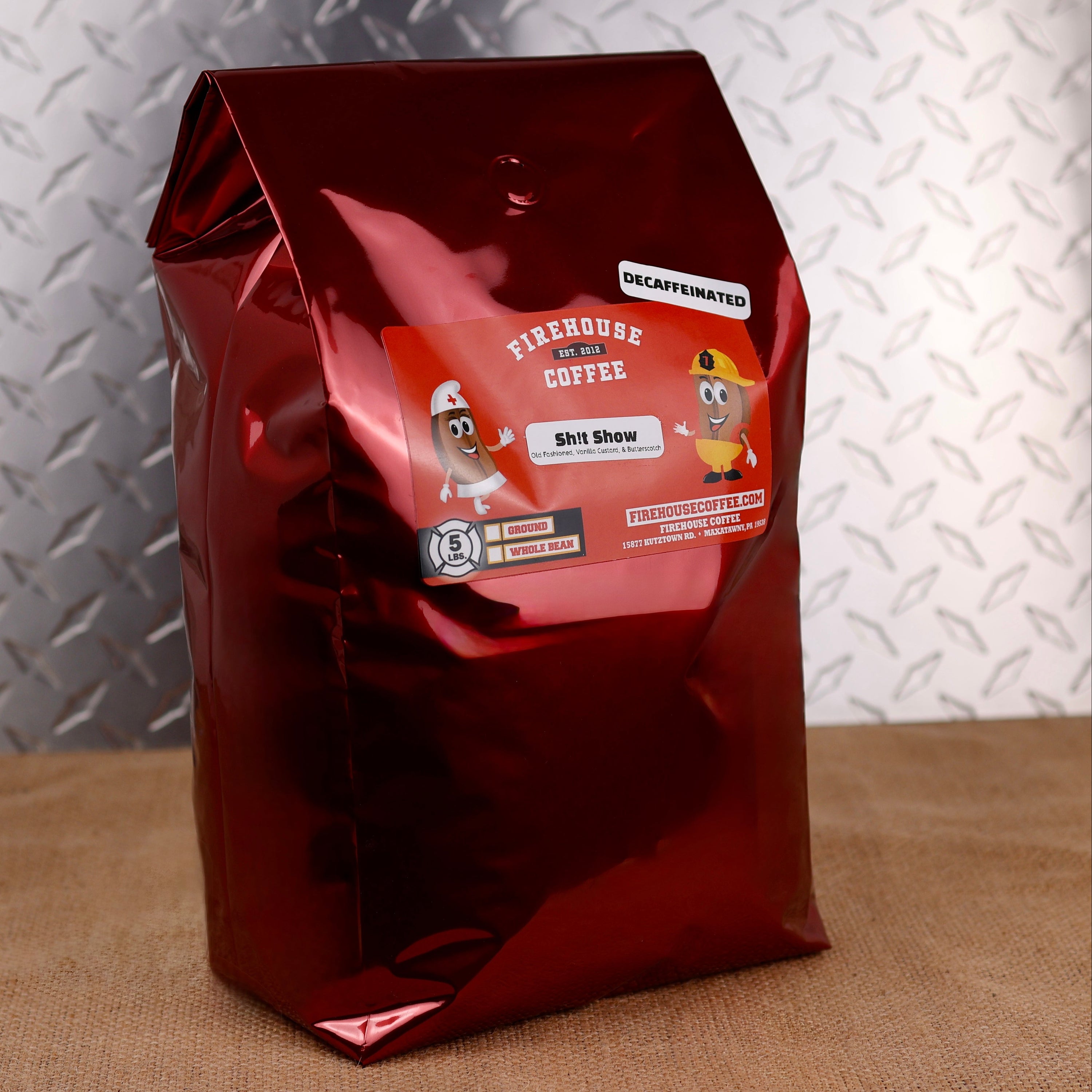 A black 12 oz Firehouse Coffee bag labeled "Shit Show" decaffeinated coffee stands in front of a matching patterned bag. The flavor profile is Old Fashioned Whiskey, Vanilla Custard, and Butterscotch, with options for ground or whole bean coffee.
