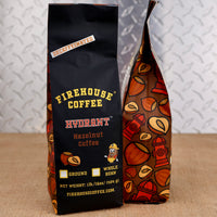 Two bags of Firehouse Coffee Hydrant Hazelnut Coffee standing on a burlap surface against a textured gray background. The left bag is solid black with yellow and red text, and the right bag has a brown base with orange, red, and yellow coffee bean illustrations.