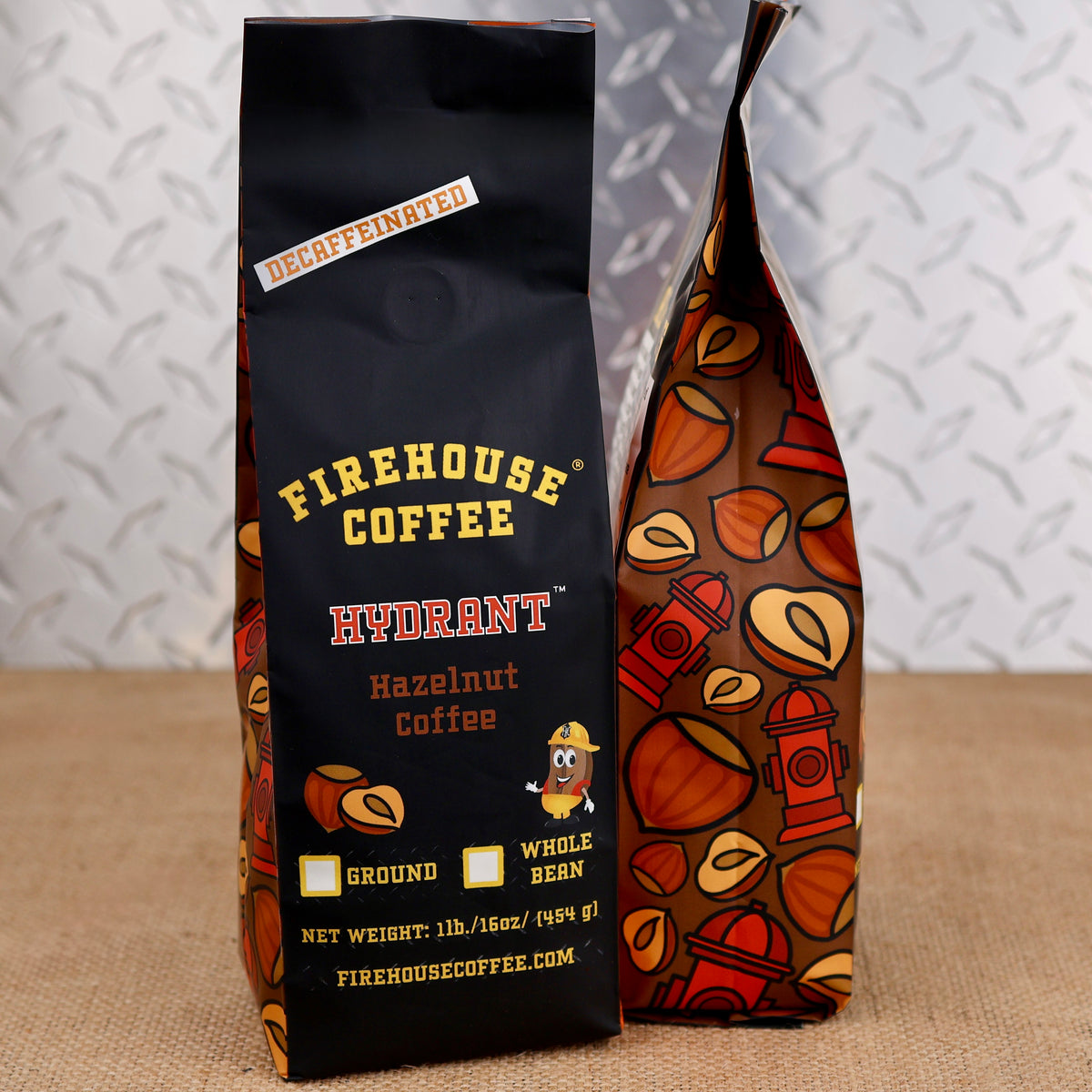 Two bags of Firehouse Coffee Hydrant Hazelnut Coffee standing on a burlap surface against a textured gray background. The left bag is solid black with yellow and red text, and the right bag has a brown base with orange, red, and yellow coffee bean illustrations.