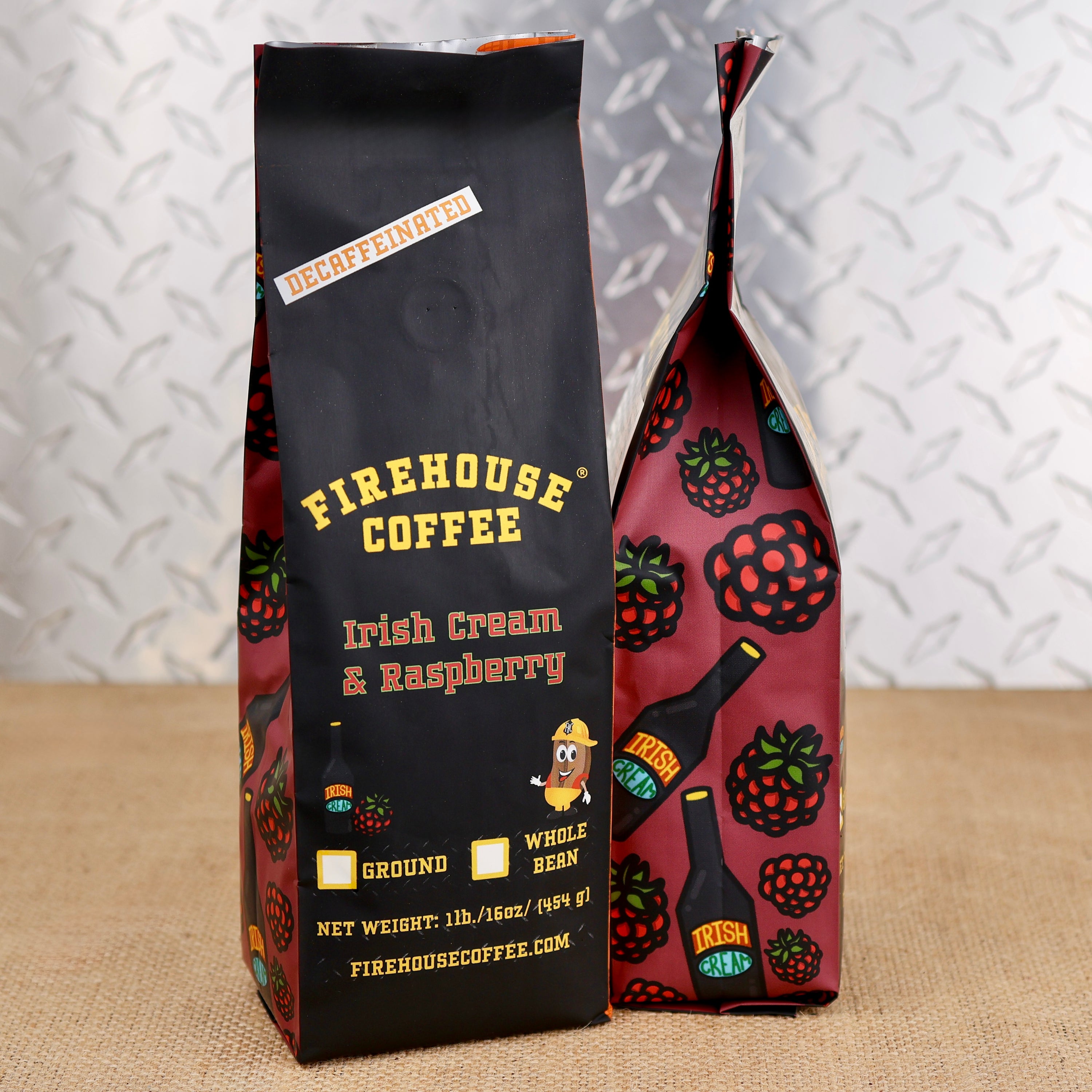Two bags of Firehouse Coffee Irish Cream & Raspberry coffee standing on a burlap surface against a diamond plate metal background. The bag in front is matte black with yellow and brown text, and the bag behind it has a raspberry and coffee bottle pattern on a dark red background.