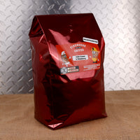 A glossy metallic dark red coffee bag standing upright on a burlap surface against a diamond-plate metal wall. The front label reads 'Firehouse Coffee Co' and 'Decaffeinated Utah Cream & Raspberry'.