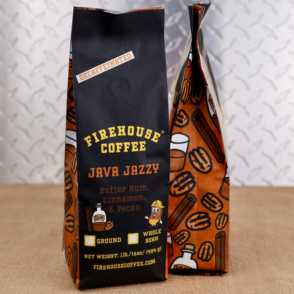 Two bags of Firehouse Coffee 'Java Jazzy' whole bean coffee standing on a burlap surface against a white diamond-plate metal background. The front bag is black with yellow and orange text and graphics, and the second bag behind it is orange with a pattern of coffee cups, cinnamon sticks, and nuts.