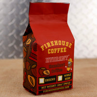 Firehouse Coffee Hydrant Hazelnut Coffee in a 12 oz red coffee bag featuring roasted hazelnut flavor, Firehouse Coffee branding, hazelnut graphics, and options for ground or whole bean coffee.