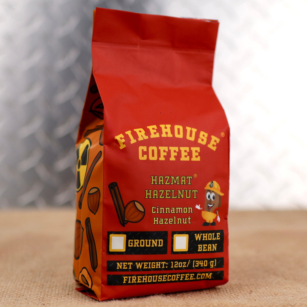 Firehouse Coffee Cinnamon Hazelnut and Hazmat Hazelnut® coffee bags, 12oz packaging featuring bold red front label, ground and whole bean options, with orange hazard symbol patterned bag in background