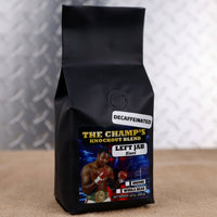 Black coffee bag labeled 'The Champ's Knockout Decaf Blend' on a textured surface with a patterned wall in the background.