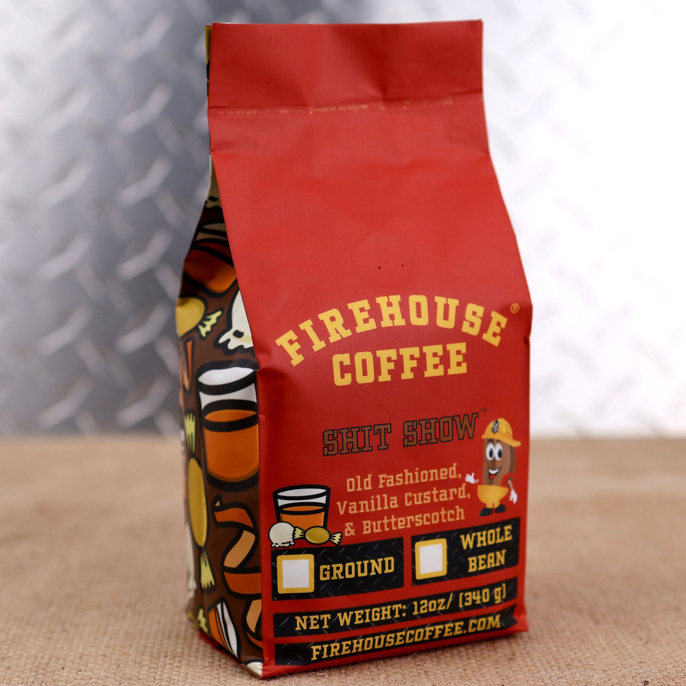 A red 12-ounce bag of Firehouse Coffee Shit Show flavored coffee stands on a burlap-covered surface against a soft gray patterned background. The front label features the Firehouse Coffee logo above the product name “Shit Show.” Flavor notes include Old Fashioned Whiskey, Vanilla Custard, and Butterscotch, illustrated with candy graphics. The label also includes the Firehouse coffee bean mascot wearing a firefighter helmet, checkboxes for ground or whole bean coffee, the net weight of 12 ounces (340 grams)